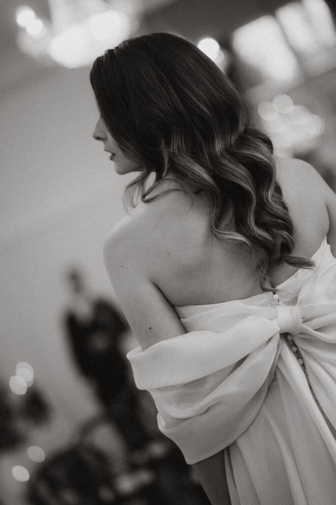 Braut von hinten mit eleganten Wellen und großer Schleife am Rückenausschnitt – Beleza Weddings & Events